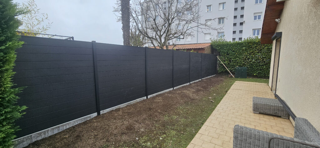 Longue clôture composite anthracite en lattes pour brise-vue de jardin, installée près d'une terrasse pavée et de terre fraîchement labourée.