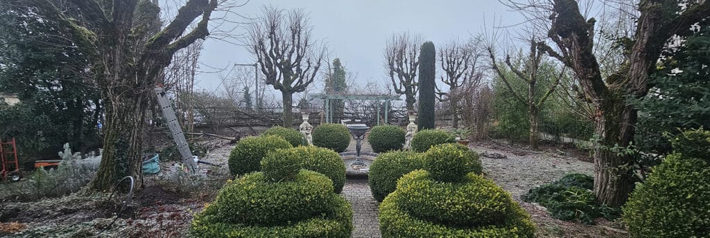 Allée pavée d'un jardin formel d'hiver. Buis taillés, fontaine centrale, statues, et grands arbres étêtés sous un ciel gris.
