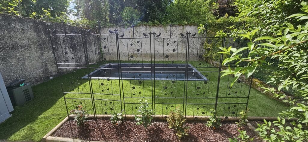 Treillage fer forgé : idée d'aménagement petit jardin. Treillage décoratif en fer forgé noir, bassin de jardin sur gazon synthétique, entouré de murs. Rosiers et paillis de gravier rouge.