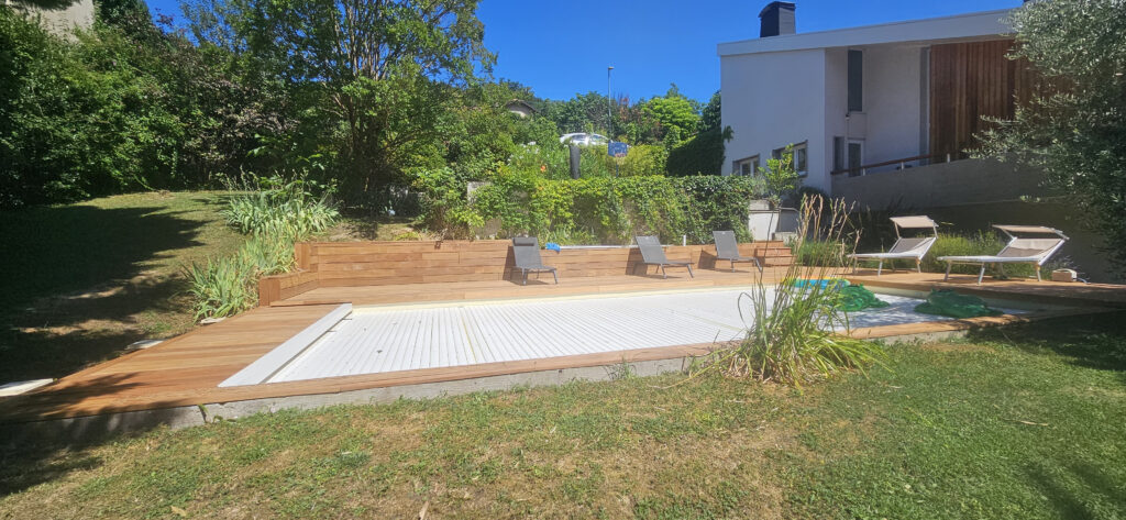 Piscine couverte à lames blanches intégrée à une grande terrasse en bois exotique. Transats modernes et mur de soutènement dans un jardin en pente.